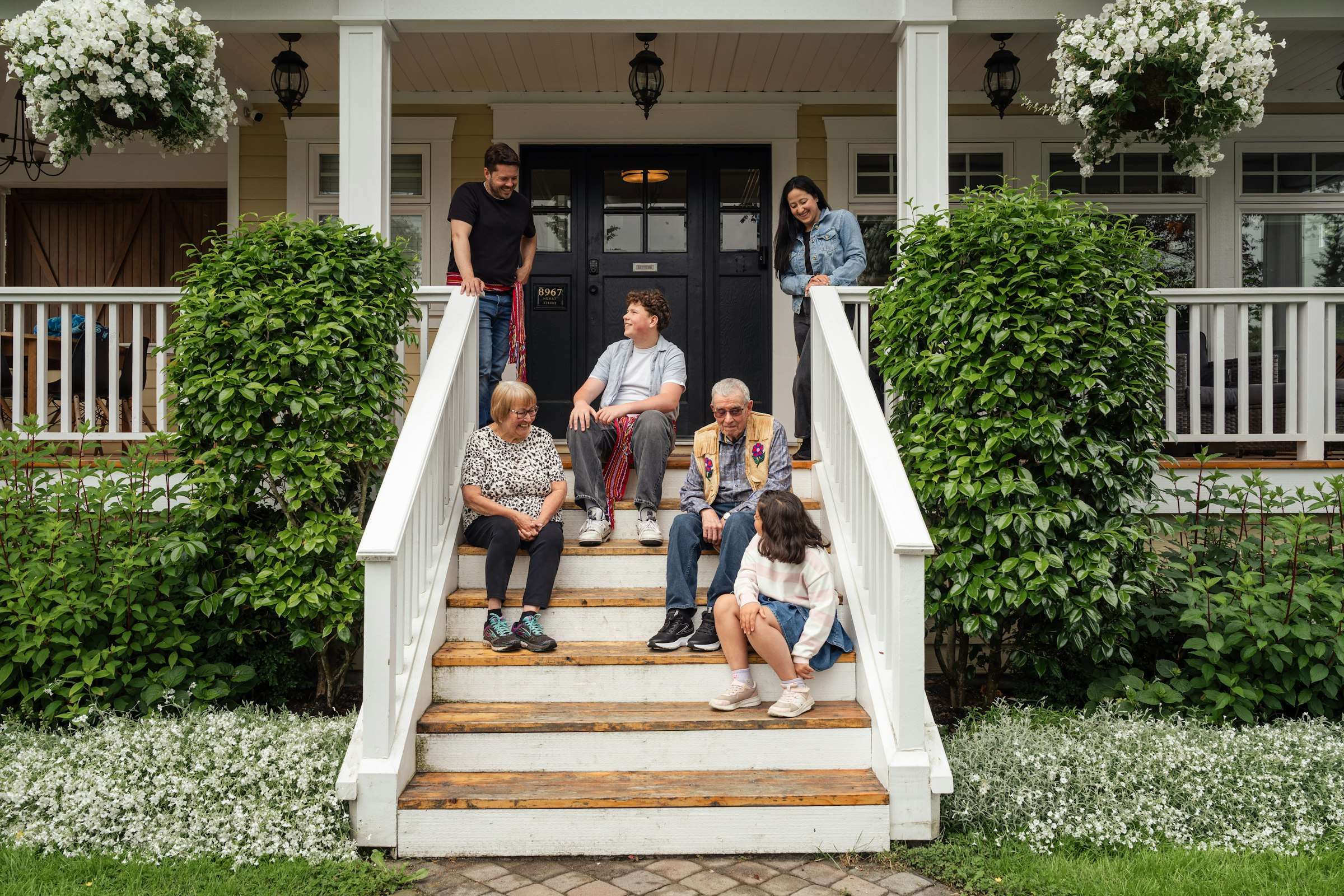 Three generations of a family sitting together on the front steps of a white house with a covered porch